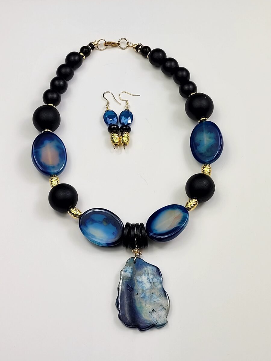 Matte black beaded necklace with glossy navy-blue agate oval accents and a large blue agate slab pendant, with crystal drop earrings.